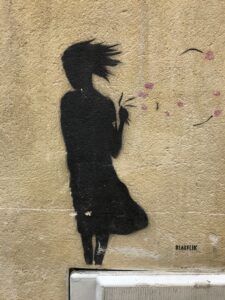 Graffiti of a silhouette of a person leaning into a wind, their hair and skirt blowing back, along with petals from a flower they're holding